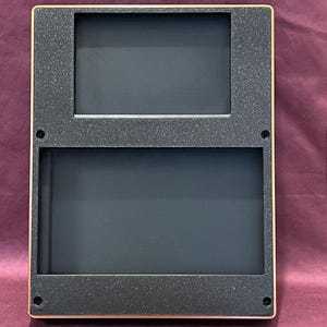May include: A rectangular, two-section frame with a dark gray textured surface and a gold-colored border. The frame has two rectangular openings, one above the other, with a dark interior. The frame is likely for displaying photos or artwork.