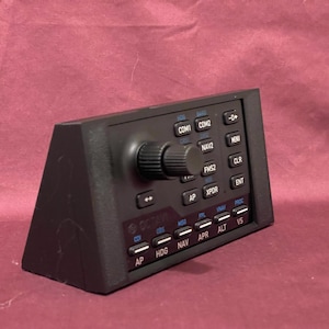 May include: A black avionics control panel with multiple buttons and a central knob. The panel has blue and white text labels, including "COM1," "COM2," "NAV2," and "FMS2." The device is angled and appears to be made of plastic.