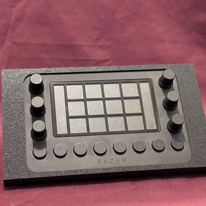 May include: Black Razer controller with a grid of square buttons and several round knobs. The controller has the Razer logo and a textured surface.