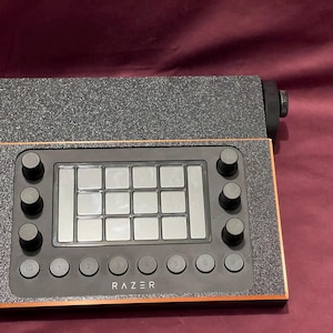 May include: A black and gray Razer audio controller with multiple buttons and pads. The controller has a rectangular design with an orange border and the Razer logo at the bottom. The top section is a textured gray.