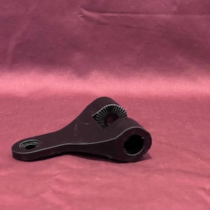 May include: A dark brown, mechanical-looking object with a curved shape and a circular opening. It has a toothed wheel-like feature and a hole at the end of a long arm. The object is set against a maroon background.