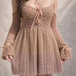 Könnte beinhalten: Beigefarbenes Häkelkleid mit langen Ärmeln und ausgestelltem Rock. Das Kleid hat schulterfreie Ärmel, ein geschnürtes Oberteil und Schleifen an den Ärmeln. Das Kleid ist handgefertigt und für besondere Anlässe geeignet.