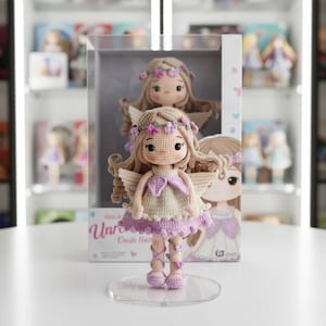 May include: A crochet doll with angel wings, a floral crown, and a lavender dress. The doll is displayed on a clear stand in front of a box with a similar doll illustration. The doll's hair is light brown, and the dress has a beige top and a lavender skirt.