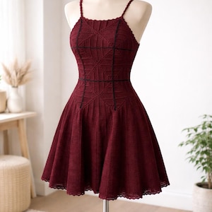 Peut inclure: Robe en crochet bordeaux sur mannequin. La robe a un corsage ajusté avec un col carré et de fines bretelles. Des lignes noires accentuent le design du corsage. La jupe s'évase à partir de la taille, et l'ourlet a un bord festonné. La robe mesure environ 80 cm de long.