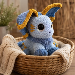 May include: A plush stuffed animal crafted from light blue, dark blue, and mustard yellow yarn. The creature features large black eyes, long floppy ears with yellow accents, and a textured, fuzzy appearance. It sits inside a woven basket.