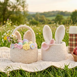 May include: Two crochet Easter baskets with bunny ears. One basket holds pastel eggs and flowers, the other has bunny ears and a pink bow. A plaid thermos and a basket of apples are also visible.