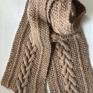 May include: A tan-colored, knitted scarf with a cable knit design. The scarf is made of a soft, textured yarn and features a thick, braided pattern running down the center. The scarf is folded over itself.
