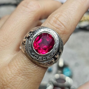 May include: A silver-toned ring with a large, oval, ruby-red gemstone. The ring has a detailed design around the gem, with the word "FORCE" visible. The ring is worn on a finger.