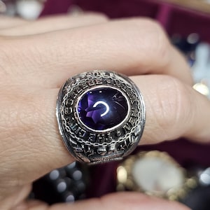 May include: A silver-toned ring with a large, oval, deep purple gemstone. The ring has intricate detailing around the band, with text and patterns. The ring is a classic style, likely a class ring or a similar commemorative piece.