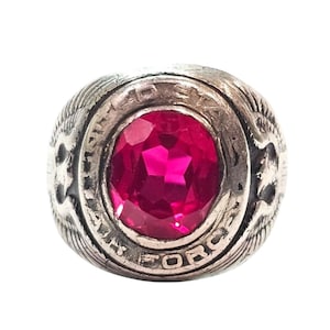 May include: A silver-toned ring with a large, oval, magenta-colored gemstone at its center. The ring's band features the words "AIR FORCE" and decorative details. The ring is likely a military or commemorative item.