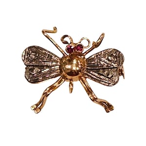 Puede incluir: Broche de insecto dorado con ojos rojos y alas detalladas. El broche tiene un cuerpo texturizado y delicadas patas y antenas. Las alas presentan un patrón geométrico. Esta joya es un accesorio único.