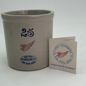 May include: A light gray stoneware crock featuring the number 25 in blue, a red wing graphic, and a blue oval stamp reading Red Wing Stoneware Co. Red Wing, Minn. Next to it is a folded paper tag with the same logo and text Continuing the Tradition.