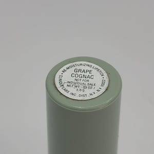 May include: A light green cylindrical lipstick tube with a silver label on the base. The label reads: RE-MOISTURIZING LIPSTICK, GRAPE COGNAC, NOT FOR INDIVIDUAL SALE, NET WT .13 OZ. / 3.6 g, CLINIQUE LABS, INC., DIST., N.Y., N.Y. 10022.