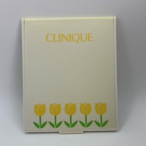 May include: A rectangular, off-white compact mirror featuring the word CLINIQUE printed in yellow at the top. The bottom section displays a row of five yellow tulips with green stems and leaves, creating a simple, clean, and functional design.