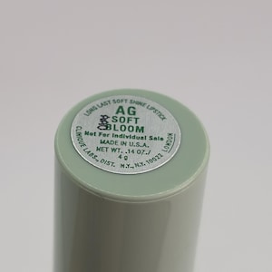 May include: A cylindrical light green lipstick tube with a silver circular label on the top. The label reads LONG LAST SOFT SHINE LIPSTICK, AG SOFT BLOOM, Not For Individual Sale, MADE IN U.S.A., NET WT. .14 OZ./4 g, CLINIQUE LABS., DIST. N.Y., N.Y. 10022 LONDON.
