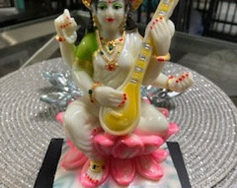 May include: A white figurine of the Hindu goddess Saraswati, seated on a pink lotus flower. She is playing a yellow veena, a stringed instrument, and is adorned with gold and green jewelry.