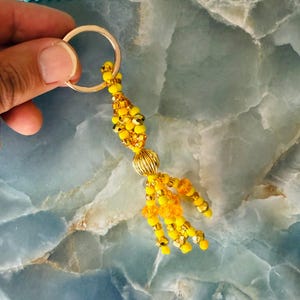 May include: A handmade beaded keyring with a gold-coloured ring. The keyring features a cluster of yellow and gold beads, with a decorative gold-coloured bead in the centre. The beads are arranged in a tassel-like design.