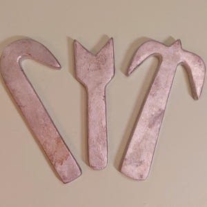 Op de afbeelding: Drie vintage metalen gereedschappen met een versleten, zilveren afwerking. De gereedschappen omvatten een gebogen haak, een gevorkt gereedschap en een hamerachtig gereedschap. De gereedschappen zijn gerangschikt op een lichtbeige oppervlak.