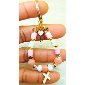 May include: A gold-colored keychain with a beaded charm. The charm features pink, white, and gold beads, a small gold star, and a white cross pendant. A gold heart and key charm hangs from the top.