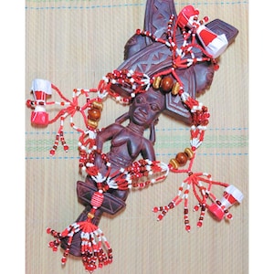 May include: A carved wooden sculpture featuring a female figure and a cross-shaped element. The sculpture is adorned with red, white, and brown beaded strands and decorative elements. The piece is set against a woven background.
