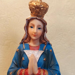 May include: A religious figurine of a woman with blue eyes, wearing a blue robe with gold trim and star accents. A gold crown with a cross sits atop her head. Her hands are clasped in prayer.
