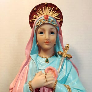 May include: A religious statue of a woman in blue robes and a pink veil. The statue features a gold crown, necklace, and bracelets. A gold sword with red accents is positioned on the side. A halo with gold rays and red accents is behind the head.