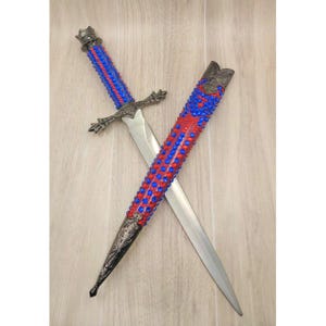 Könnte beinhalten: Ein dekorativer Dolch mit silberner Klinge und kunstvollem Knauf. Der Griff ist mit roten und blauen Perlen umwickelt, und die Scheide ist ähnlich verziert. Der Dolch und die Scheide werden auf einer hellen Holzoberfläche präsentiert.