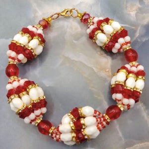 May include: A beaded bracelet with alternating sections of red and white beads. The bracelet has gold-coloured accents and a gold clasp. The design incorporates small red and white beads, larger red beads, and white, oval-shaped beads.