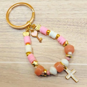 May include: A beaded keyring with pink, white, and gold beads. The keyring features a gold-coloured ring, heart charms, a key charm, and a white cross charm. The beads are arranged in a bracelet-like design.