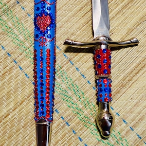 May include: A decorative dagger and sheath set. The blue sheath and handle are adorned with red and blue jewels. The dagger has a silver blade and guard. The set is displayed on a woven mat.