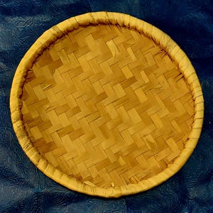 May include: A round, woven bamboo tray with a light brown color. The tray features a tightly woven pattern and a wrapped edge. The tray is likely used for serving or displaying food items.
