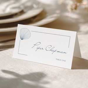 Op de afbeelding: Een witte naamkaart met een blauwe zeeschelp illustratie en een blauwe rand. De kaart leest "Piper Chapman" en "TABLE ONE" in elegant blauw schrift. Het Welcome Design Co. logo staat rechtsboven.