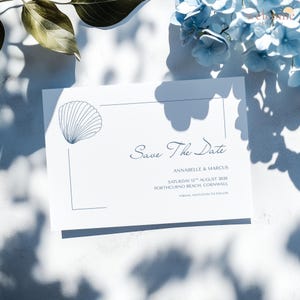 May include: A white "Save The Date" card with a seashell illustration and text in navy blue. The card includes the names Annabelle & Marcus, the date, and location: Porthcurno Beach, Cornwall. Blue flowers and shadows are in the background.