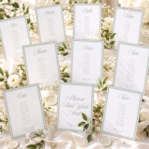 May include: A collection of ten white wedding seating chart cards with sage green borders and elegant script typography. Each card displays a table number or a welcome message, followed by a list of guest names in black ink on textured paper.