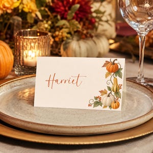 May include: A white rectangular place card featuring the name Harriet written in a rust-colored script font. The right side of the card is decorated with a botanical illustration of orange pumpkins, a yellow gourd, and autumn leaves.