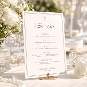 May include: A rectangular white bar menu card with a thin black border, featuring the text The Bar, sections for Cocktails, Wine, Beer, Spirits, and Soft Drinks, and the names Madeleine & Frederick with the date May 21, 20XX, held by a gold stand.