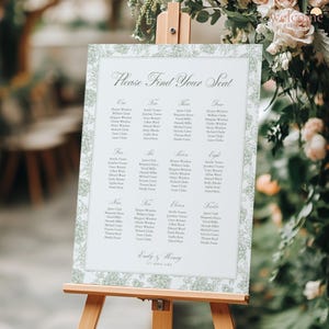 May include: A wedding seating chart on a wooden easel. The white sign has a green floral border and lists names under numbers one through twelve. The text "Please Find Your Seat" is at the top. The names are arranged in columns.
