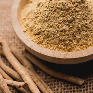 May include: A wooden bowl filled with a light brown powder sits on a textured surface. Several light brown, dried root pieces are scattered around the bowl, suggesting a natural health product or ingredient.