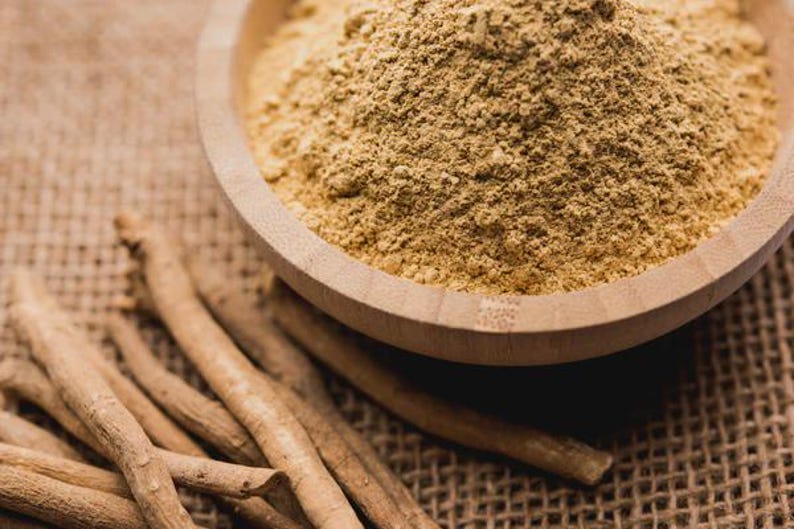 May include: A wooden bowl filled with a light brown powder sits on a textured surface. Several light brown, dried root pieces are scattered around the bowl, suggesting a natural health product or ingredient.