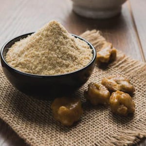 May include: A black bowl filled with a light brown powder, possibly a spice or flour, sits on a burlap cloth. Several golden-brown, irregularly shaped pieces are scattered nearby. The items are arranged on a wooden surface.