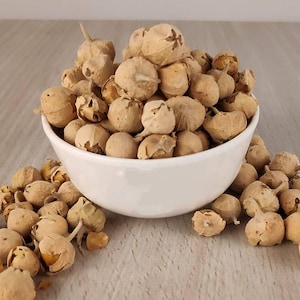 Peut inclure: Un bol en céramique blanc rempli de physalis séchés de couleur beige. Quelques fruits débordent du bol sur la surface claire. Les fruits ont une enveloppe papyracée et une forme ronde, suggérant un produit alimentaire naturel.