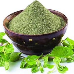 May include: A wooden bowl filled with green powder, possibly a spice or herb, sits on a white surface. Fresh green mint sprigs are arranged around the bowl. The bowl is dark brown with small, decorative gold-colored accents.