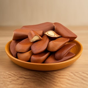 Op de afbeelding: Een houten kom gevuld met bruine noten, waarvan sommige doormidden zijn gesneden om een crèmewit interieur te onthullen. De noten hebben een gladde, langwerpige vorm en zijn gerangschikt in een warme, uitnodigende presentatie. De kom is lichtbruin.