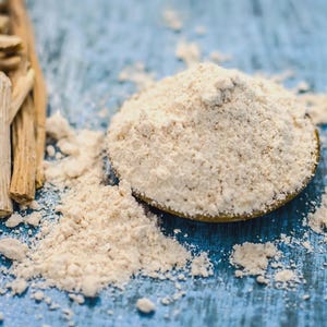 May include: Close-up of ashwagandha powder in a small bowl, with a pile of the powder scattered around it. Ashwagandha sticks are arranged to the left. The background is a blue wooden surface.