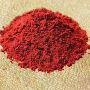 Puede incluir: Un montón de polvo rojo vibrante, posiblemente un pigmento o tinte, descansa sobre una superficie clara y texturizada. El polvo tiene una textura fina y granulada, y la imagen está bien iluminada, resaltando el color y la textura.
