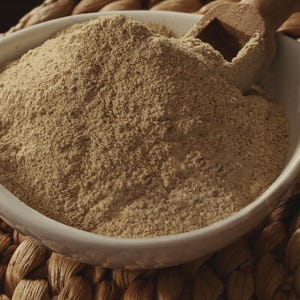 Peut inclure: Un bol en c&eacute;ramique blanc rempli d'une poudre brun clair, possiblement une &eacute;pice ou de la farine, avec une cuill&egrave;re en bois partiellement ins&eacute;r&eacute;e. Le bol repose sur une surface tiss&eacute;e et textur&eacute;e. La poudre semble finement moulue.