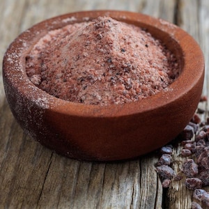 May include: A wooden bowl filled with pink Himalayan salt, with a pile of salt crystals next to it. The salt has a reddish-pink hue and a coarse texture. The bowl is brown and sits on a weathered wooden surface.
