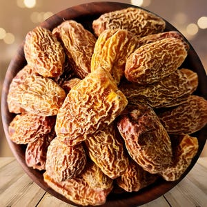 Puede incluir: Un cuenco de madera lleno de dátiles secos. Los dátiles son de color marrón claro con una textura arrugada. El cuenco es de madera marrón oscuro y está sobre una superficie de madera.