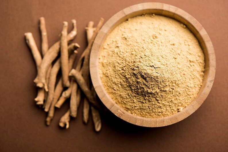 Peut inclure: Un bol en bois rempli d'une poudre brun clair est plac&eacute; &agrave; c&ocirc;t&eacute; d'un tas de racines s&eacute;ch&eacute;es de couleur beige. Les &eacute;l&eacute;ments sont dispos&eacute;s sur une surface brune. La poudre semble finement moulue et les racines ont une texture rugueuse.