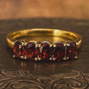 Könnte beinhalten: Ein goldener Ring mit fünf oval geschliffenen, tiefroten Edelsteinen. Der Ring liegt auf einer dekorativen Oberfläche im antiken Stil. Das Band des Rings ist goldfarben poliert. Die Edelsteine sind gleichmäßig verteilt und fangen das Licht ein.